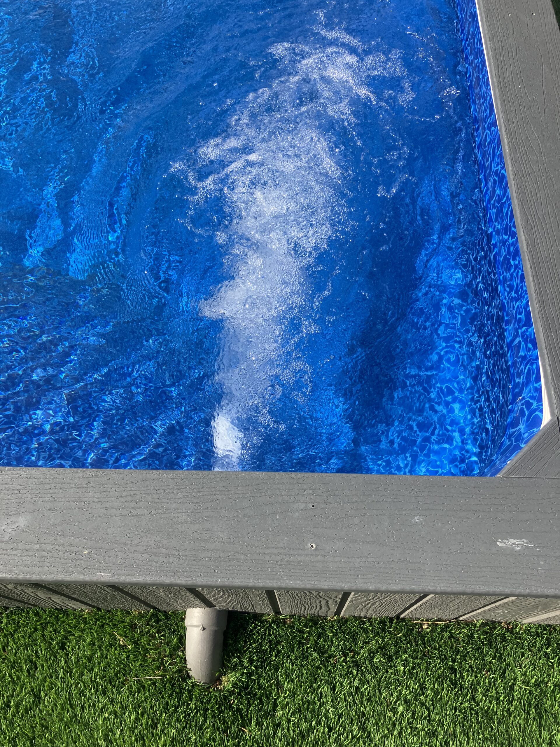 Top-down view of a bright blue rectangular swimming pool with a gray wooden deck surrounding it and green grass nearby.