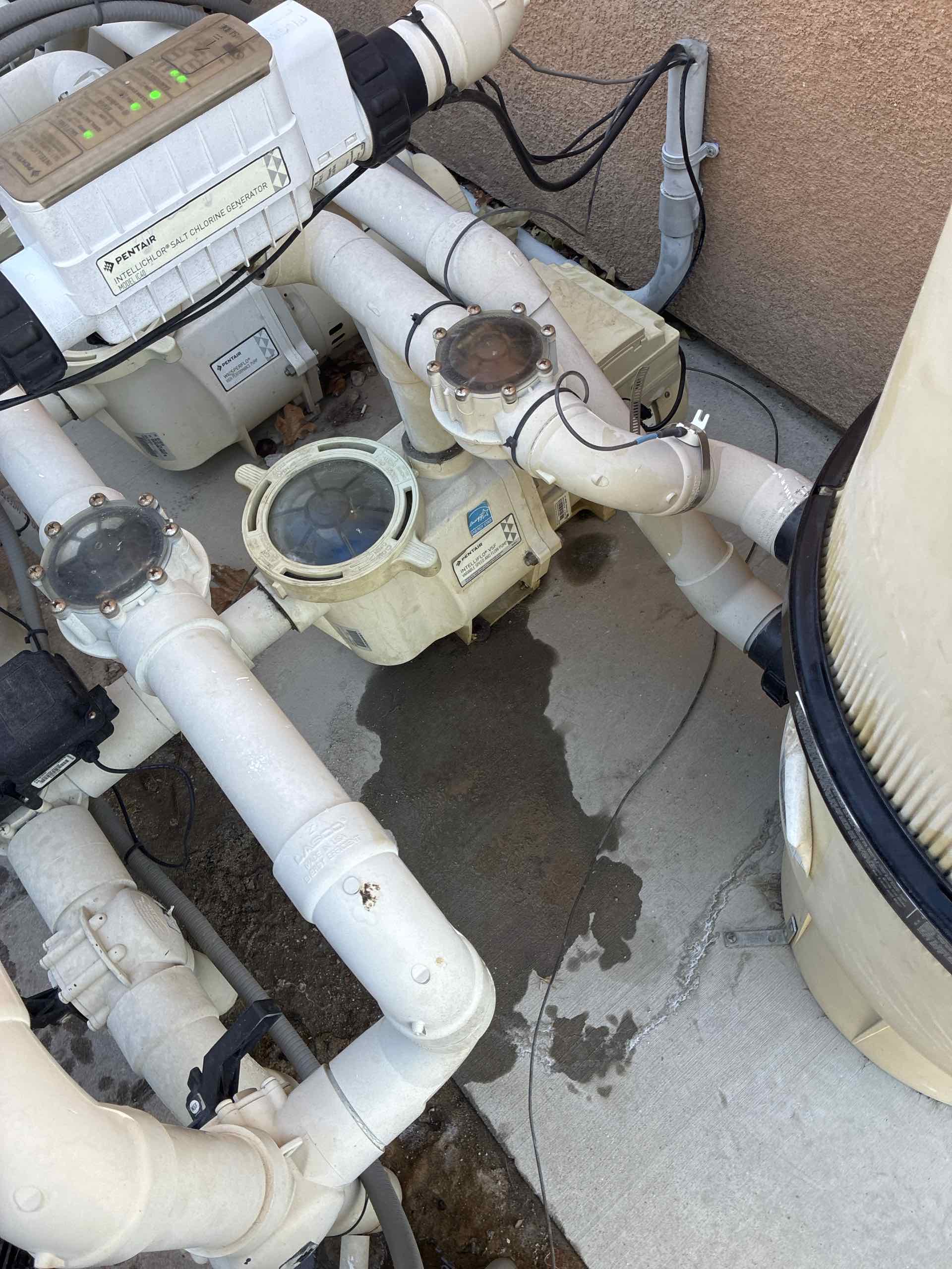 Outdoor pool equipment with white PVC pipes and a Pentair chlorine generator unit mounted on a concrete pad behind a wall.