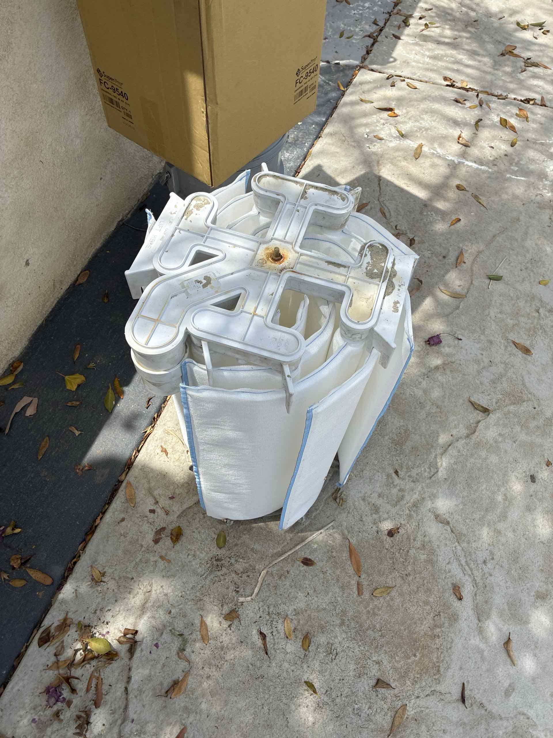 White cross-shaped plastic component sitting on top of white bags outside, with a cardboard box leaning against a wall and scattered autumn leaves on a concrete surface.