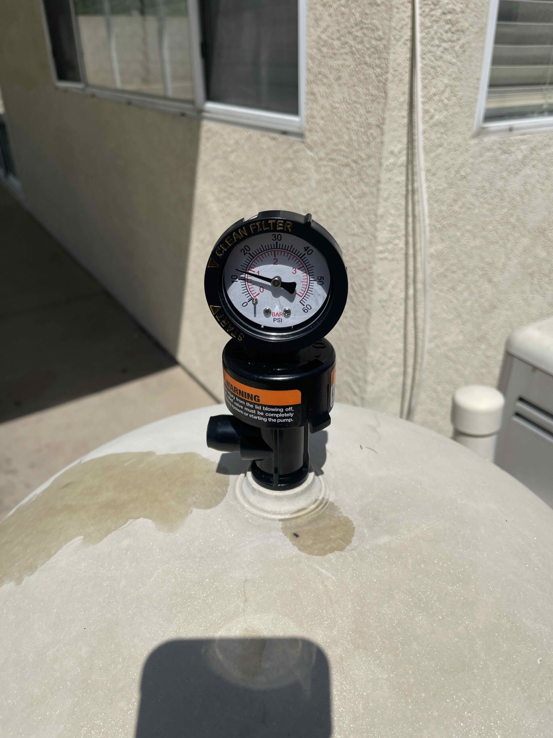 Pool filter pressure gauge labeled 'Clean Filter' mounted on a pump, with a beige textured wall behind it.