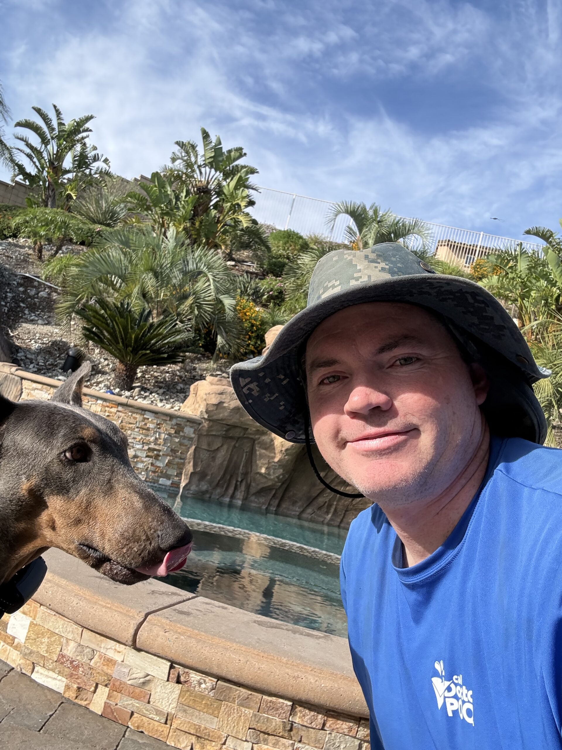 Close-up selfie of a man in a camo hat and blue shirt with a Doberman near a pool and tropical plants in the background