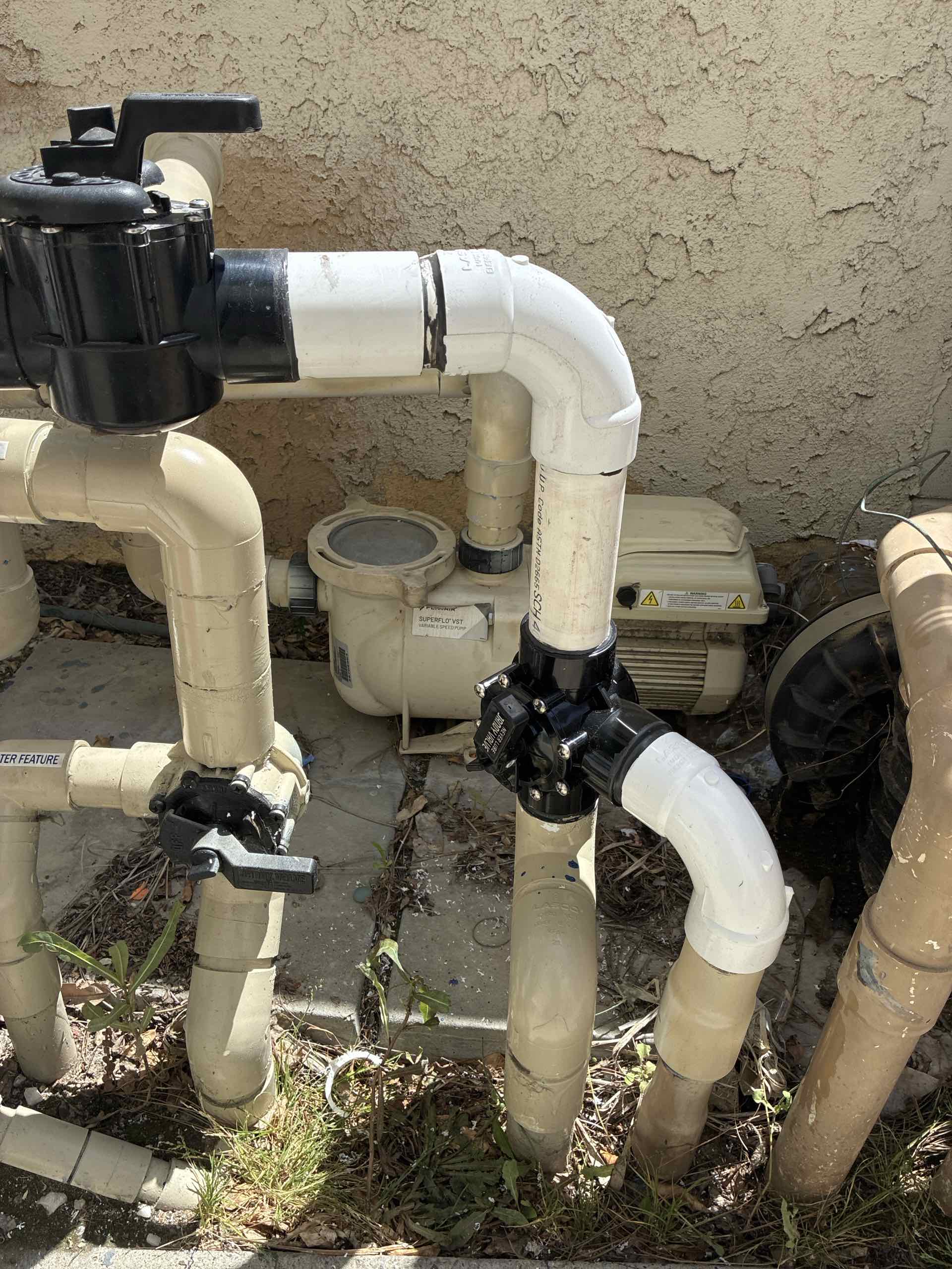Pool pump and white PVC piping with a black motor, outdoor setup against a textured beige wall.