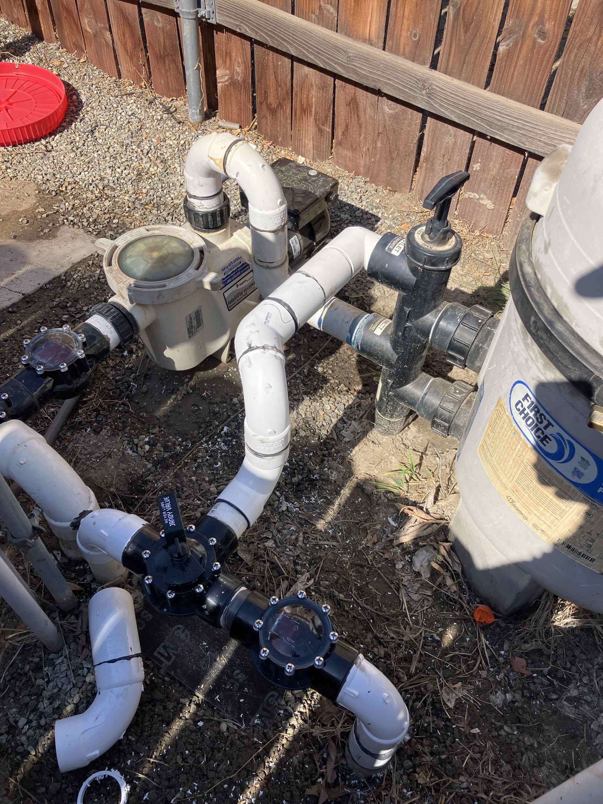 Close-up of a pool filtration system with white PVC pipes, a pump, and black valve handles outside near a wooden fence.