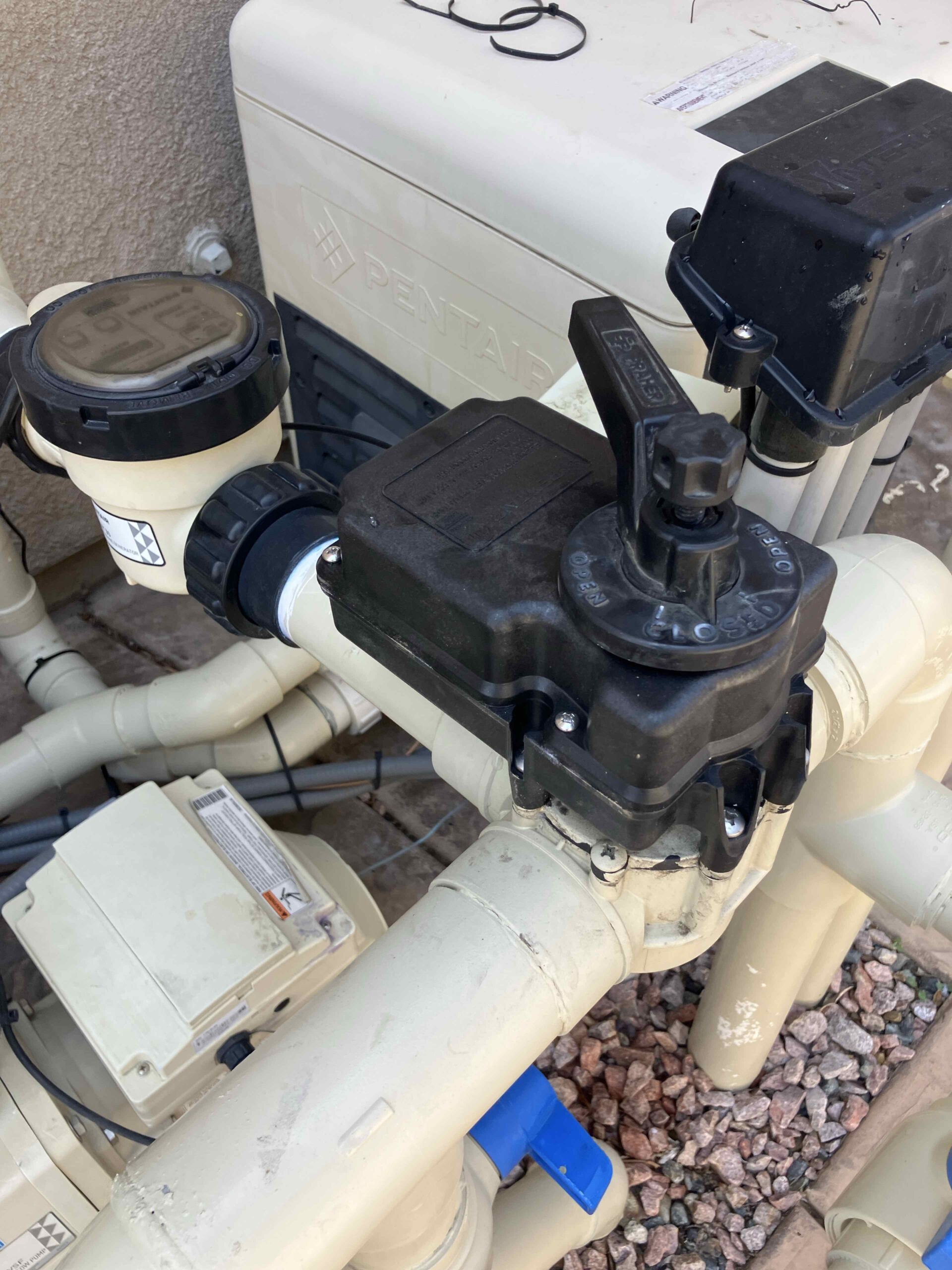 Close-up of a pool pump and filtration valves with beige PVC piping, outdoors on gravel, showing the Pentair logo on the unit.