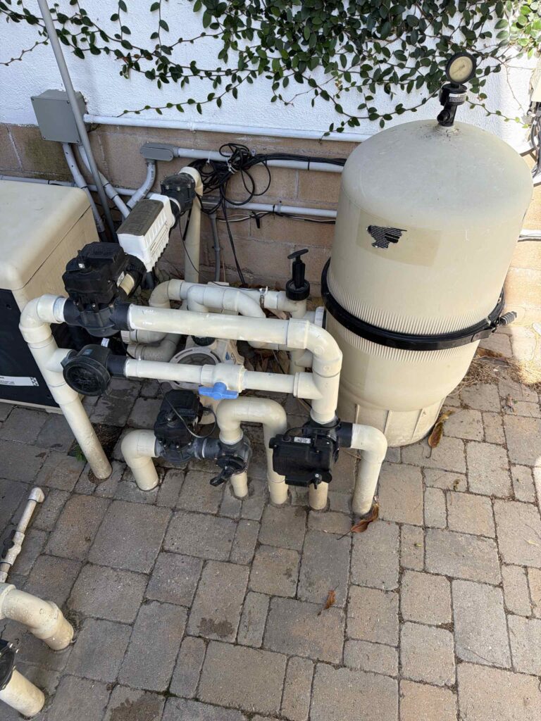 Pool equipment setup: large beige sand filter tank, pumps, and white PVC piping against a wall with climbing vines.